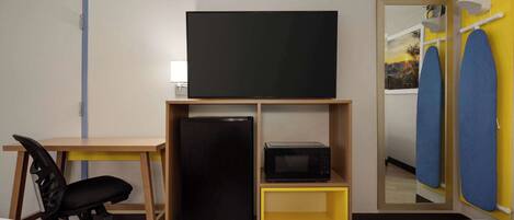Una habitación con una televisión sobre un soporte de madera, un refrigerador negro, un microondas, un escritorio con una silla y una cama con cubrecolcha de diseño geométrico.