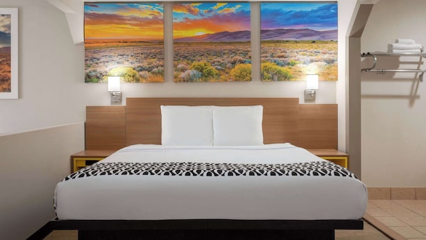 Quarto de hotel com uma cama grande, cabeceira, mesa de cabeceira, prateleira na parede e uma pintura de uma paisagem ao pôr do sol.