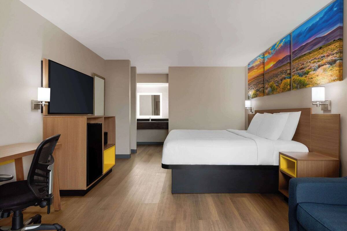 Ein Hotelzimmer mit Bett, Schreibtisch, Stuhl, Fernseher, Kunstwerken und einem Spiegel.