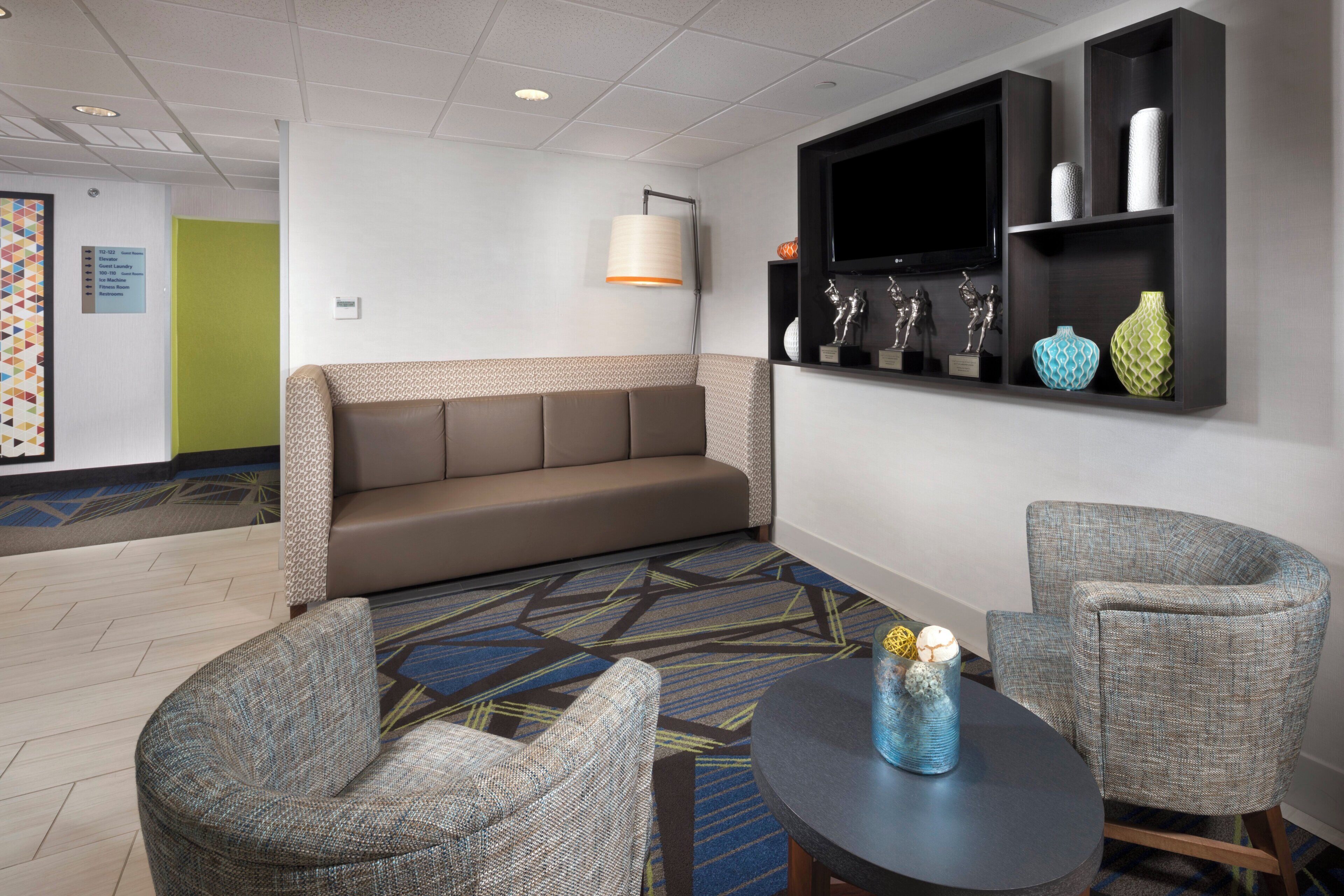 A hotel lobby with a sofa, two armchairs, a round table, a wall-mounted TV, and a shelf with decorative items.
