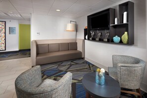A hotel lobby with a sofa, two armchairs, a round table, a wall-mounted TV, and a shelf with decorative items.
