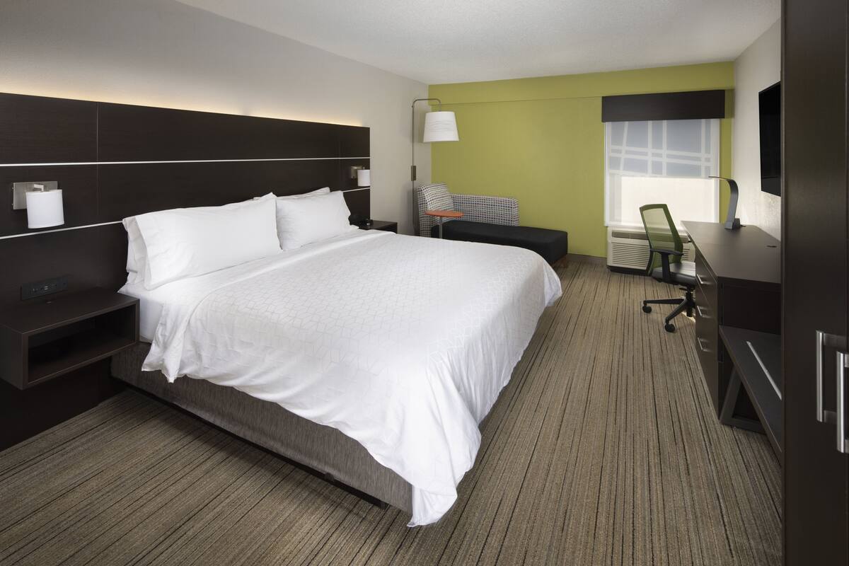 A hotel room with a large bed, a desk with a chair, a TV, and a window with blinds.