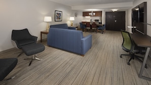 A hotel room with a blue sofa, a dark armchair, a small desk, and a dining table with chairs.