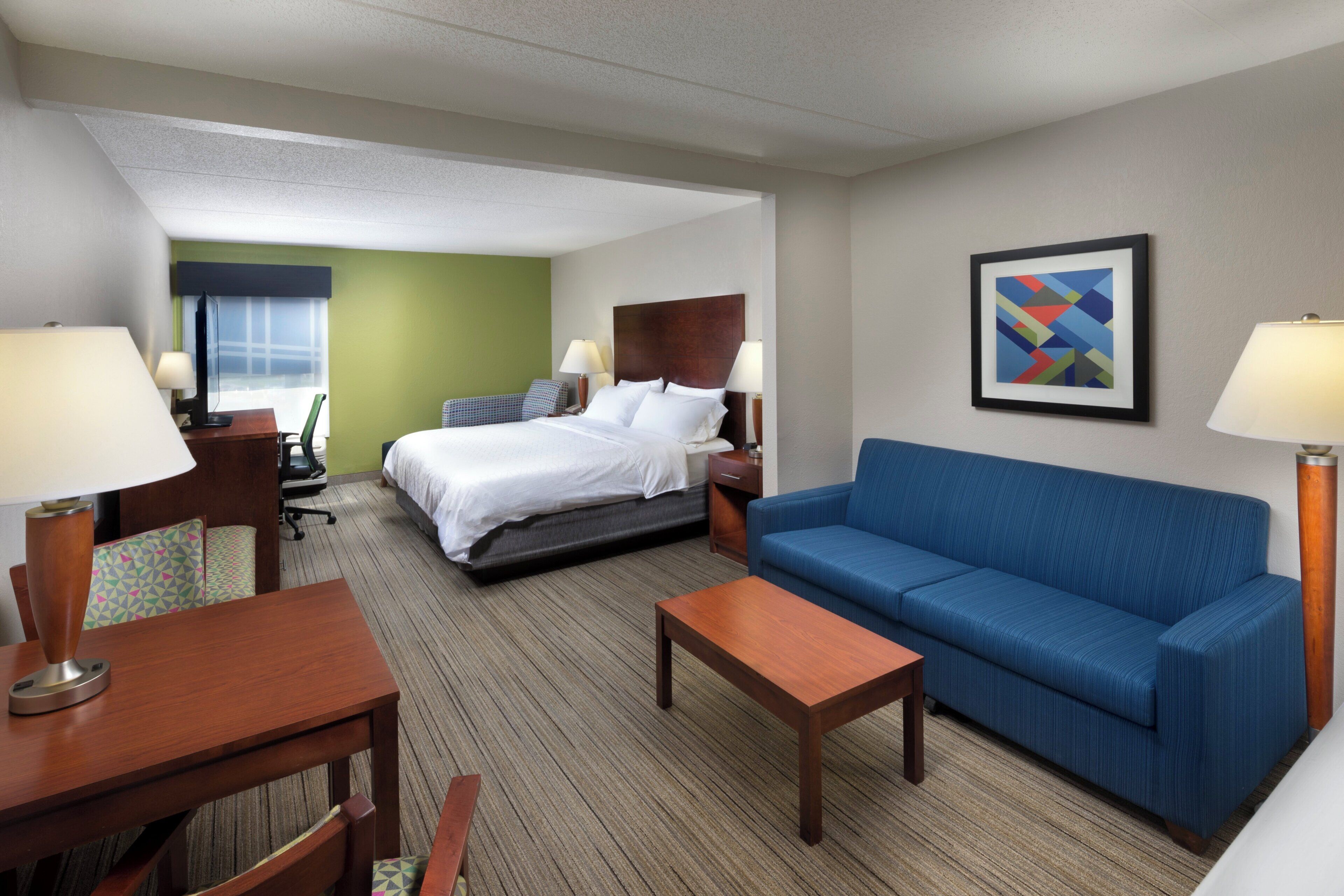 Habitación de hotel con cama, sofá azul, mesa de centro de madera, escritorio con silla y lámpara.