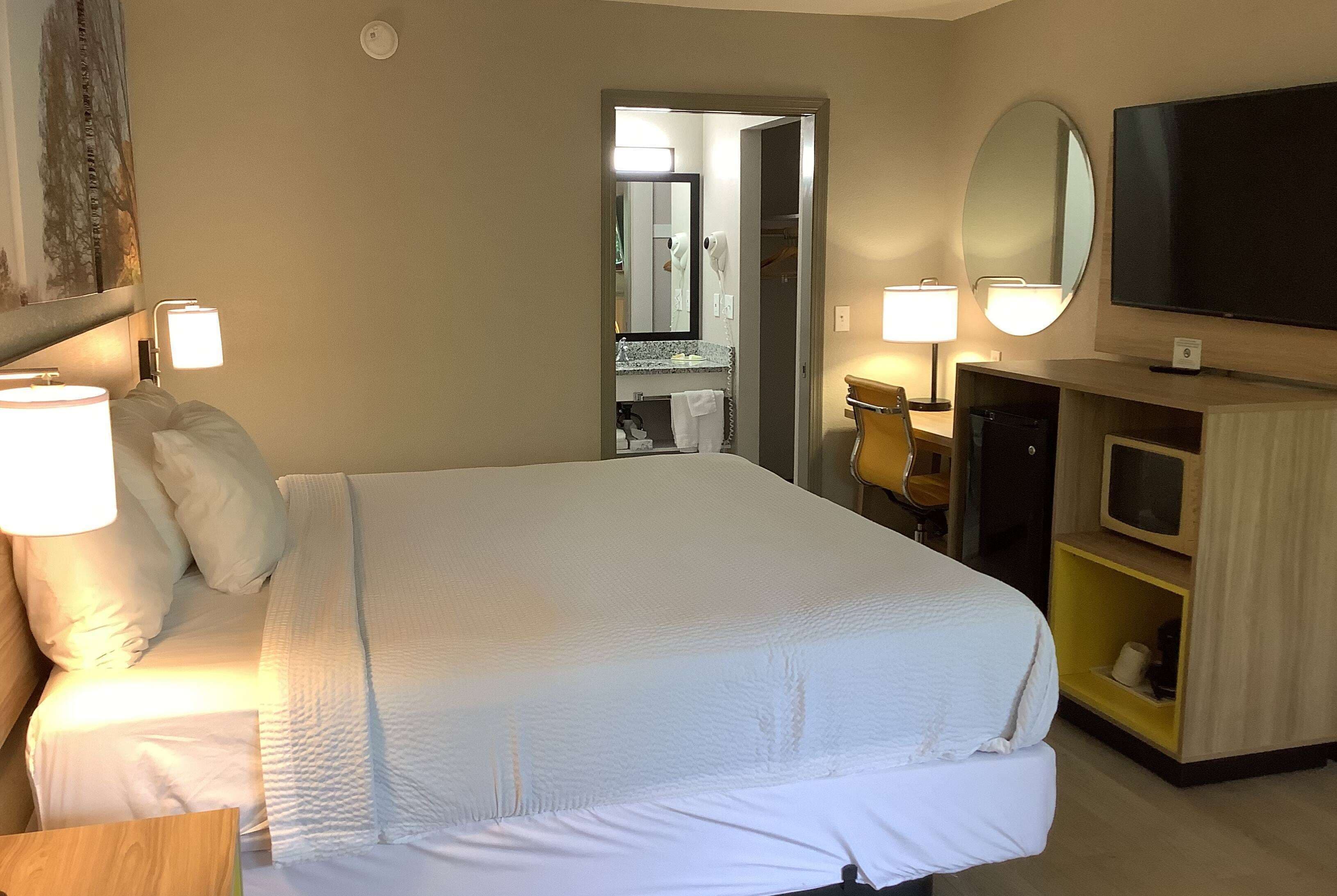 A hotel room with a large bed, bedside lamps, a TV, a mirror, and a bathroom visible through an open door.