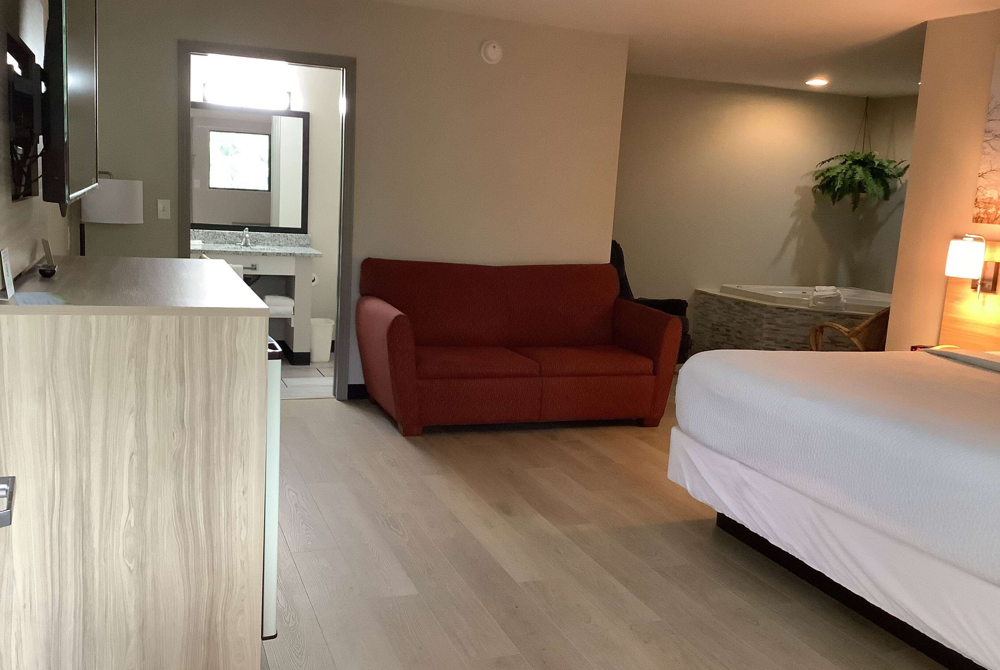 Habitación de hotel con piso de madera, un sofá rojo, una cama con ropa blanca, una mesita y un espejo sobre el sofá.