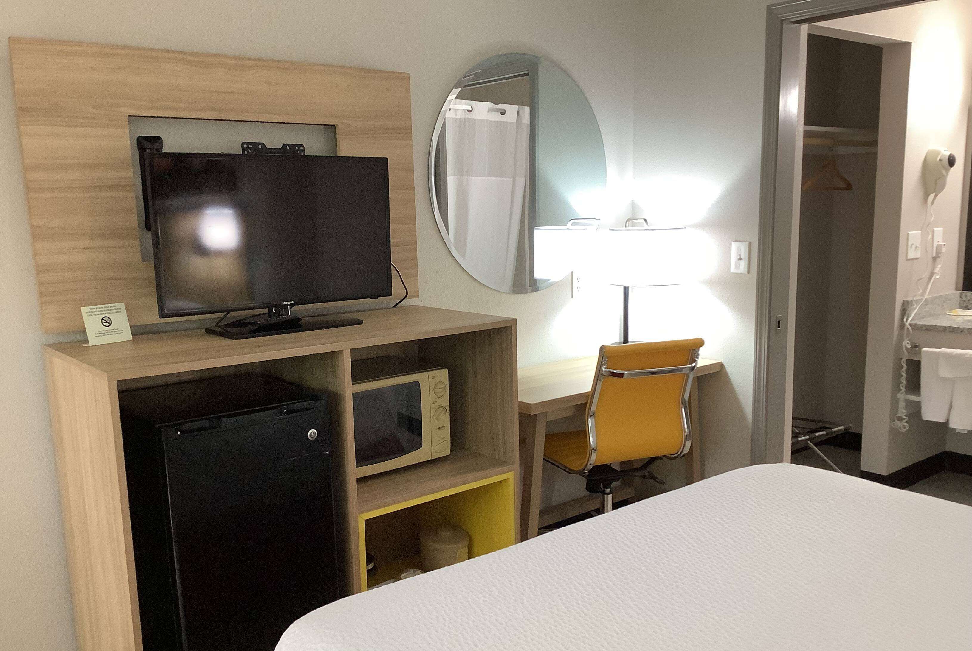 Una camera d'albergo con un letto, una scrivania con un computer, una televisione, un forno a microonde, una sedia, uno specchio e un angolo doccia.