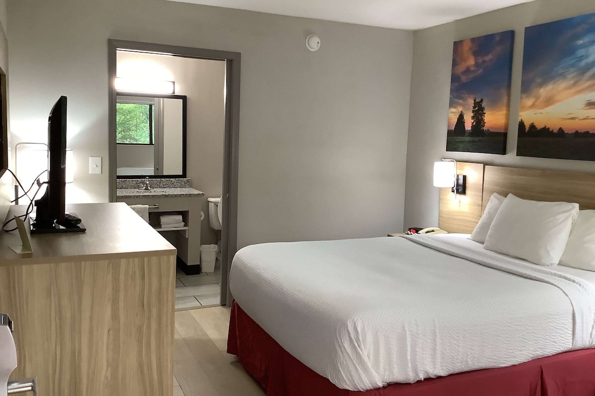 A hotel room with a bed, a desk, a mirror, a window, and two paintings on the wall.