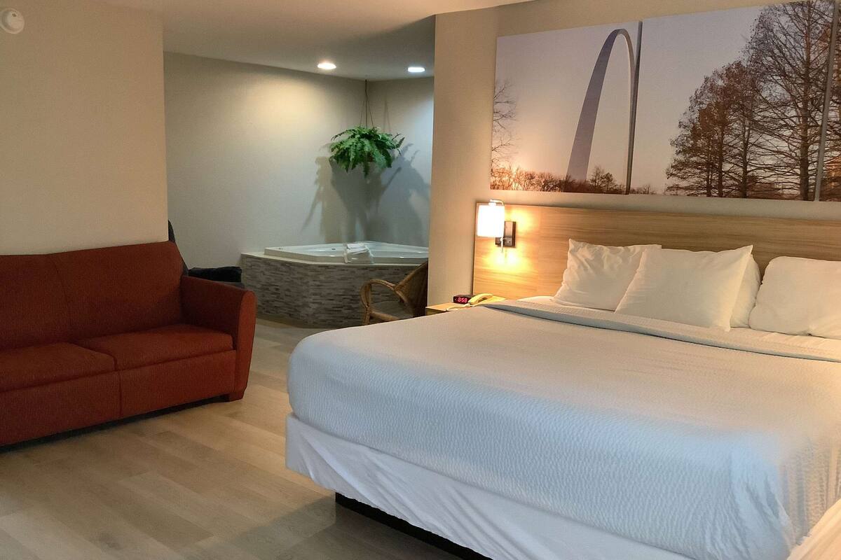 A hotel room with a large bed, a red sofa, a wall-mounted TV, and a large window with a view of trees.