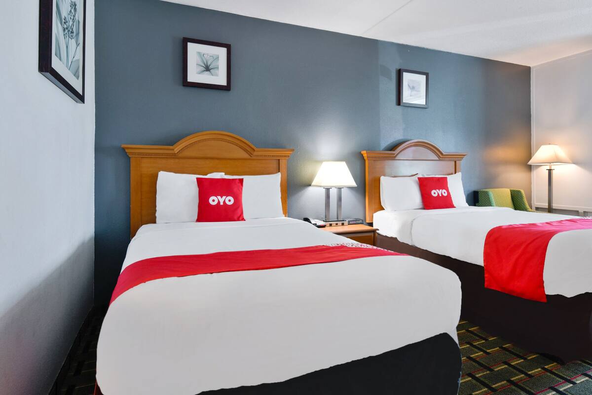 A hotel room with two beds, each with a red OYO pillow, a bedside lamp, and a framed picture on the wall.