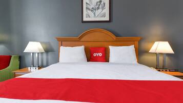Una cama bien hecha con una colcha roja y blanca, dos lámparas de noche, una silla verde y un cuadro enmarcado en la pared.