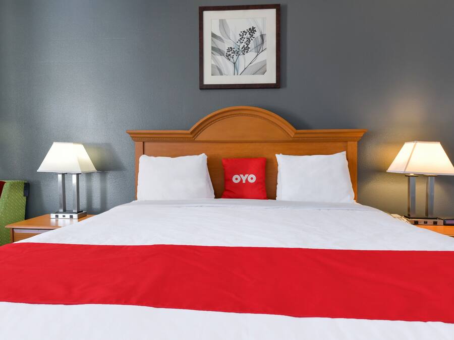 Una cama bien hecha con una colcha roja y blanca, dos lámparas de noche, una silla verde y un cuadro enmarcado en la pared.