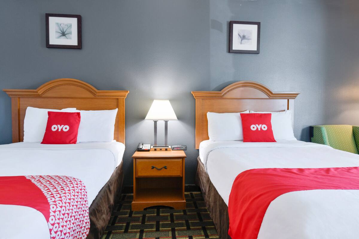 A hotel room with two beds, each with a red OYO pillow, a bedside table with a lamp, and a framed picture on the wall.