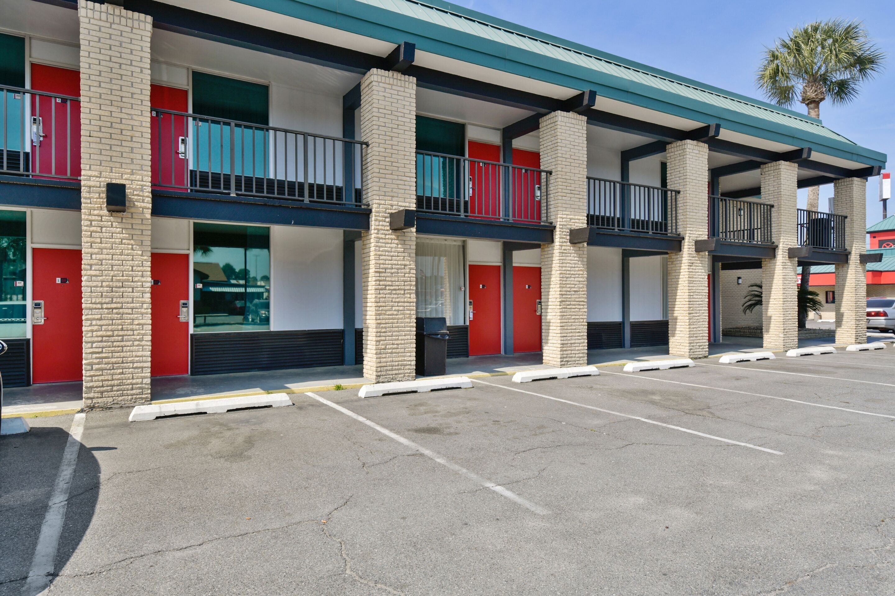 A multi-story hotel with red doors and balconies, a parking lot, and a palm tree in the background.