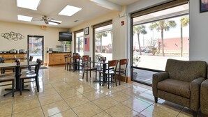 A spacious dining area with multiple tables and chairs, a kitchen area with a refrigerator and microwave, and large windows offering a view of palm trees and a parking lot. - OYO Hotel Lake Park/Valdosta I-75, Exit 5 (Lake Park)