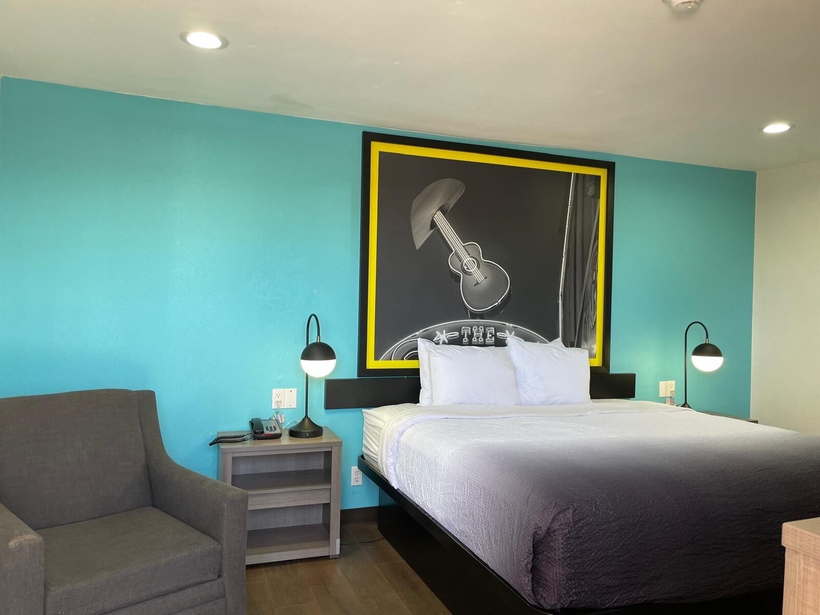 A hotel room with a bed, a chair, a nightstand, and a wall with a guitar artwork.
