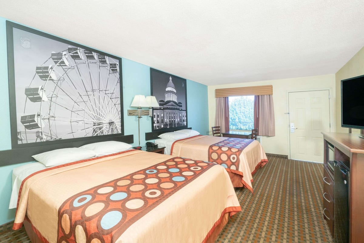 A hotel room with two beds, a large wall mural, a desk, a television, and a window with curtains.