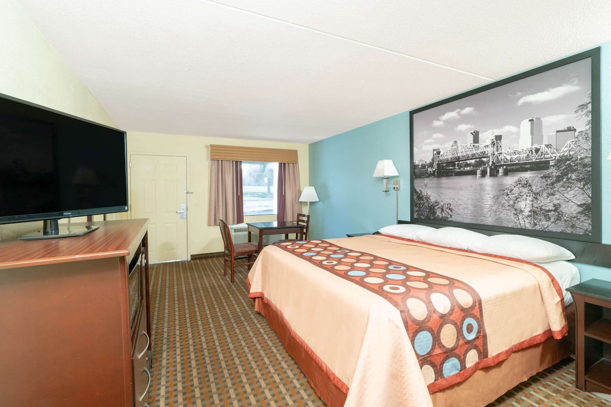 A hotel room with a bed, a desk with a TV, a chair, a lamp, and a large framed picture on the wall.