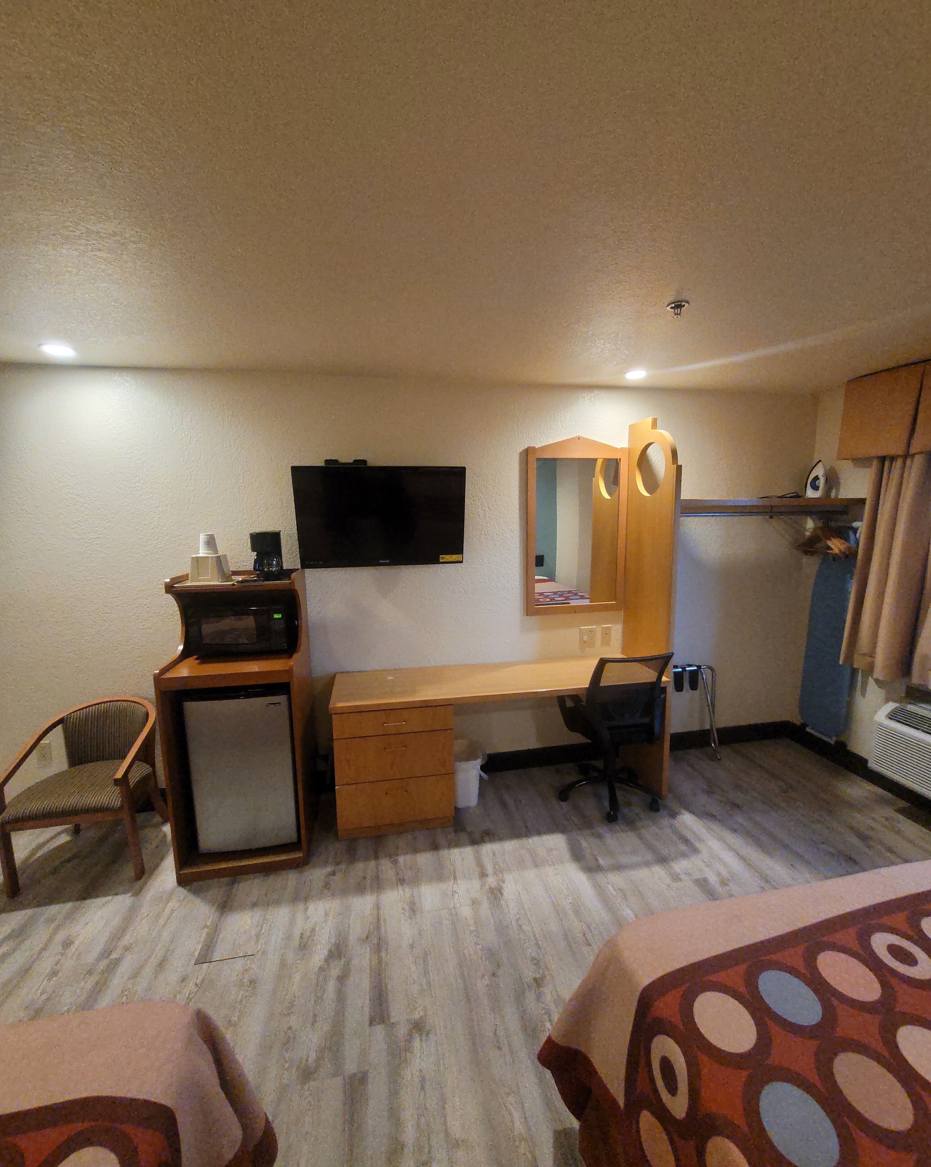 Ein kleines Hotelzimmer mit Schreibtisch aus Holz, einem Stuhl, einem Fernseher, einem Spiegel und einer kleinen Küchenzeile.