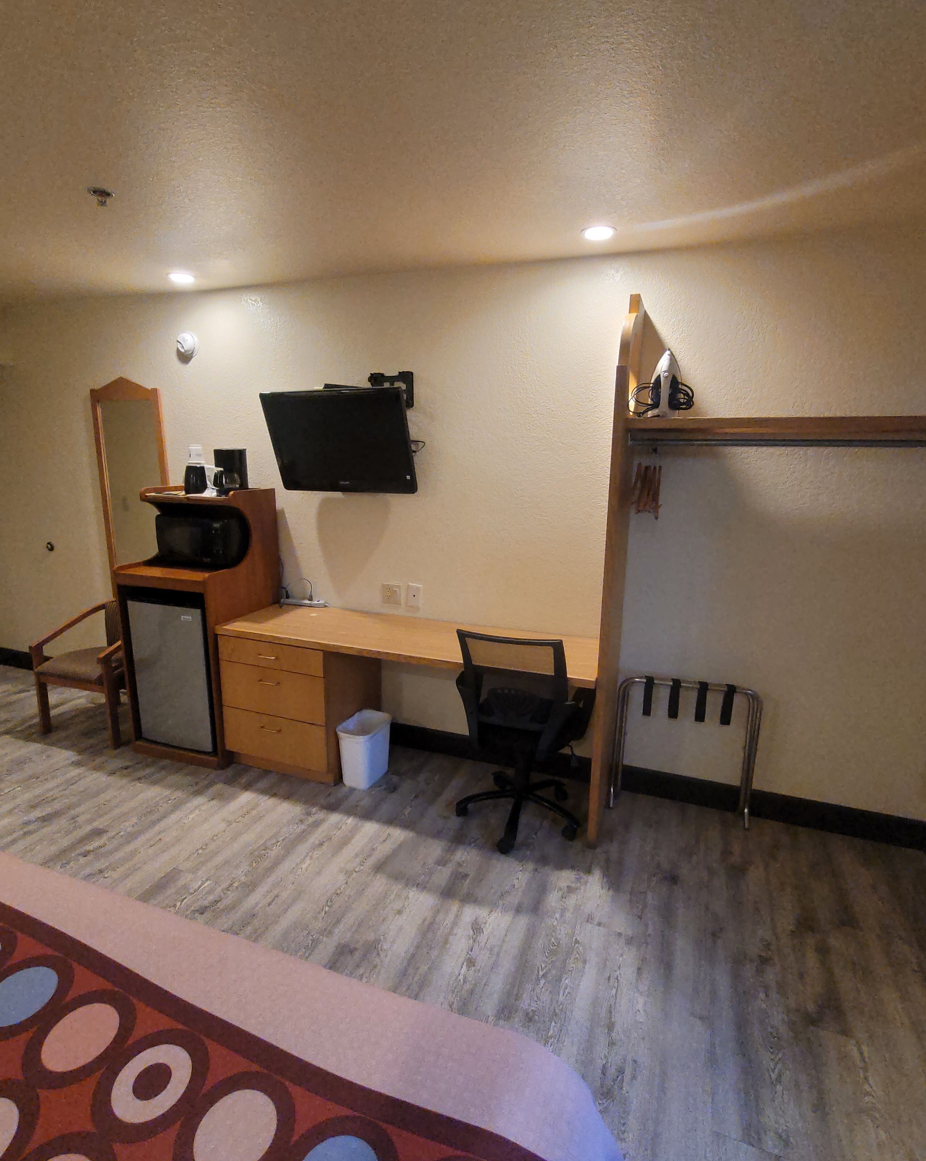 A compact hotel room with a wooden desk, a chair, a small refrigerator, a trash bin, and a television mounted on the wall.