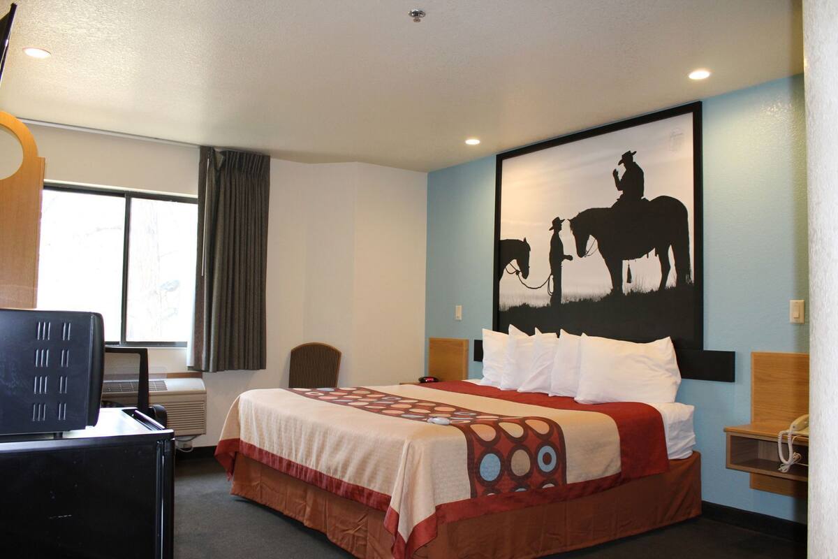 A hotel room with a bed, a desk, a chair, a window with curtains, and a wall painting.