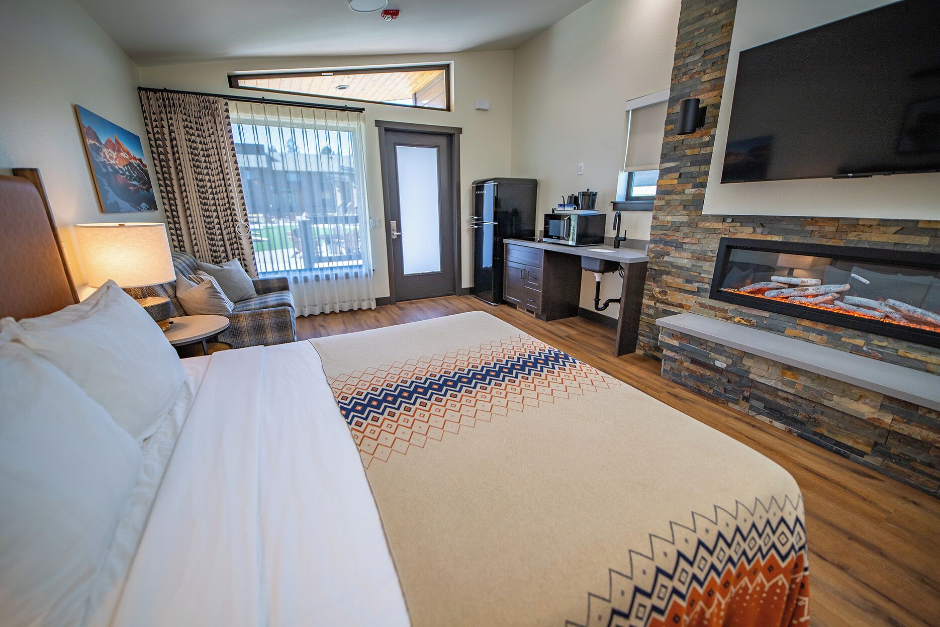 A hotel room with a bed, a desk, a TV, a fireplace, and a balcony with curtains.