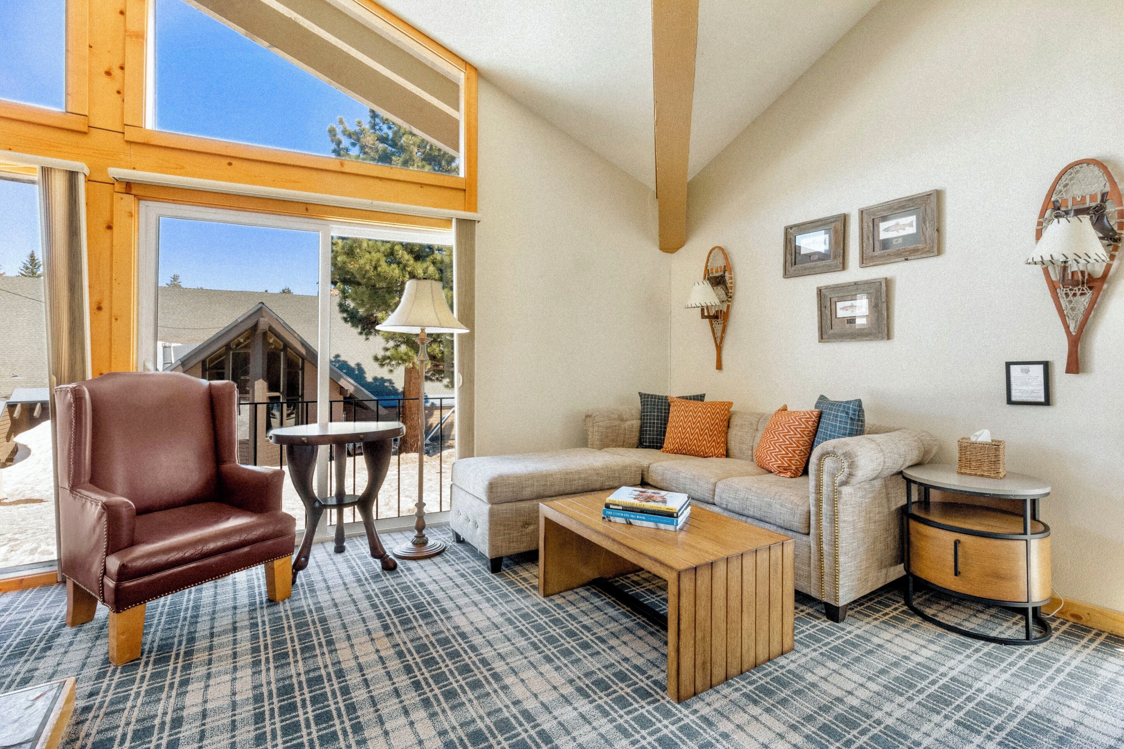 A cozy living room with a leather chair, a sofa, a coffee table, and a small side table. There are framed pictures on the wall and a window with a view of the outdoors.
