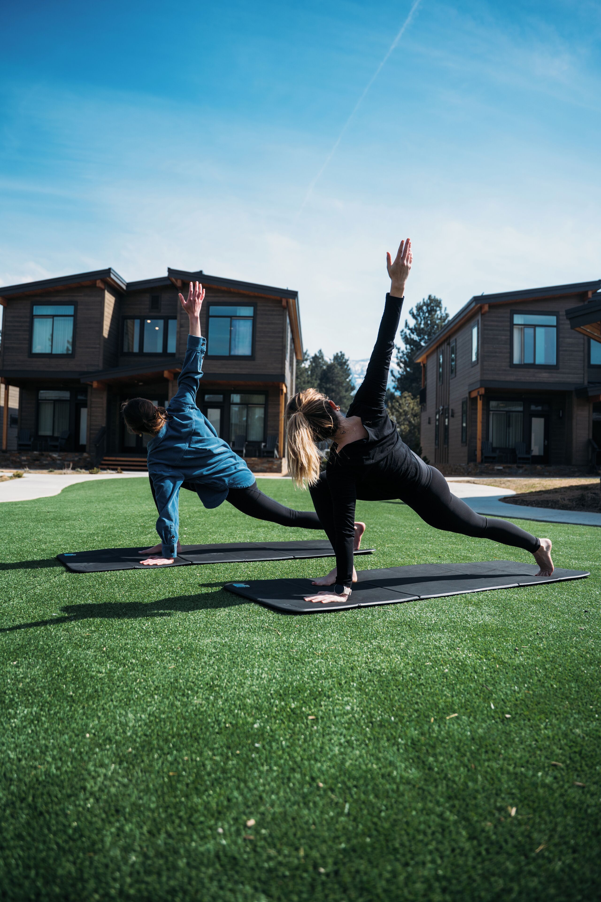 Twee personen doen yoga op buitenmatten voor een modern huis met een verzorgd gazon.