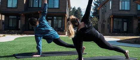 Twee personen doen yoga op buitenmatten voor een modern huis met een verzorgd gazon.