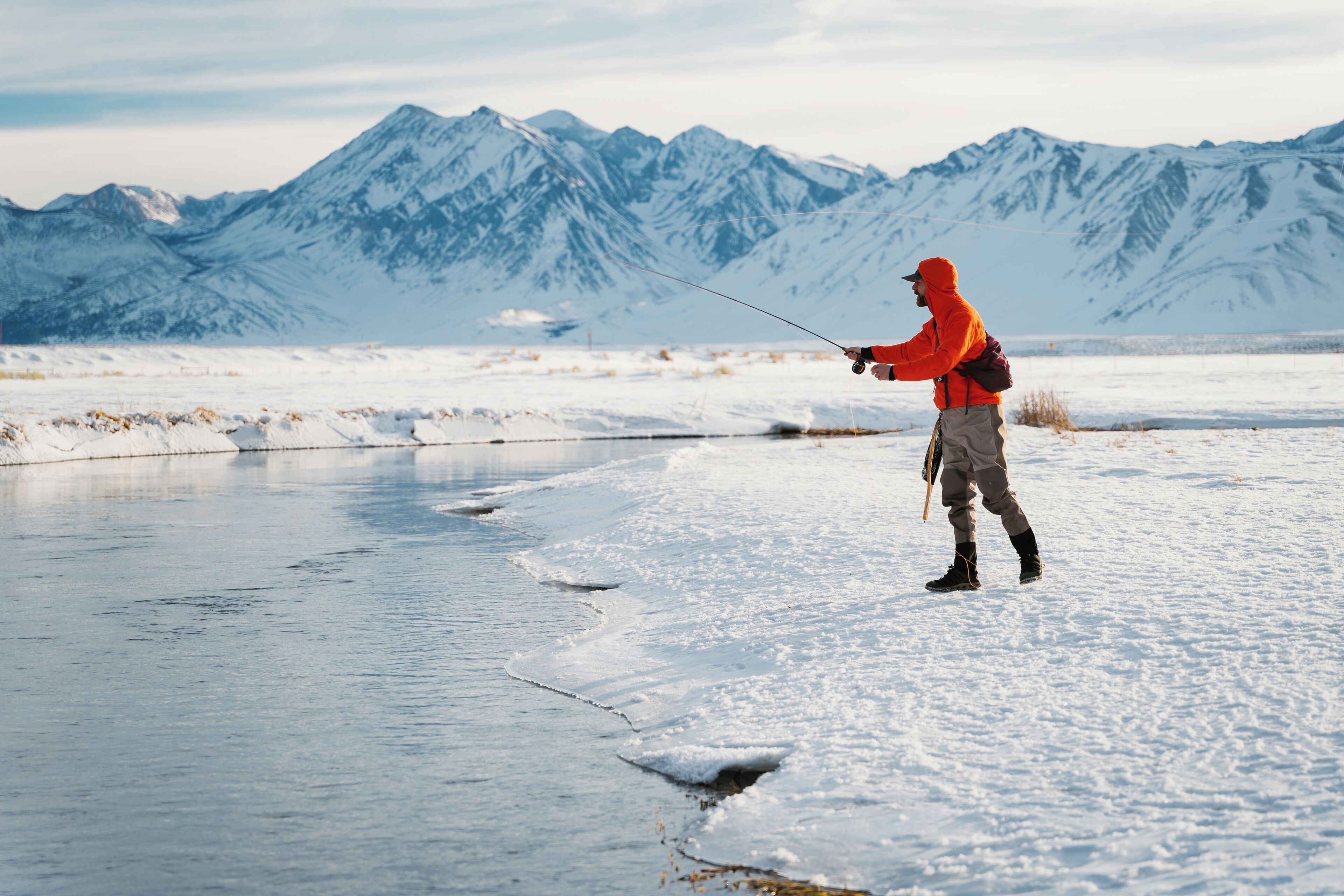 A person fishing in a snowy landscape with mountains in the background.
