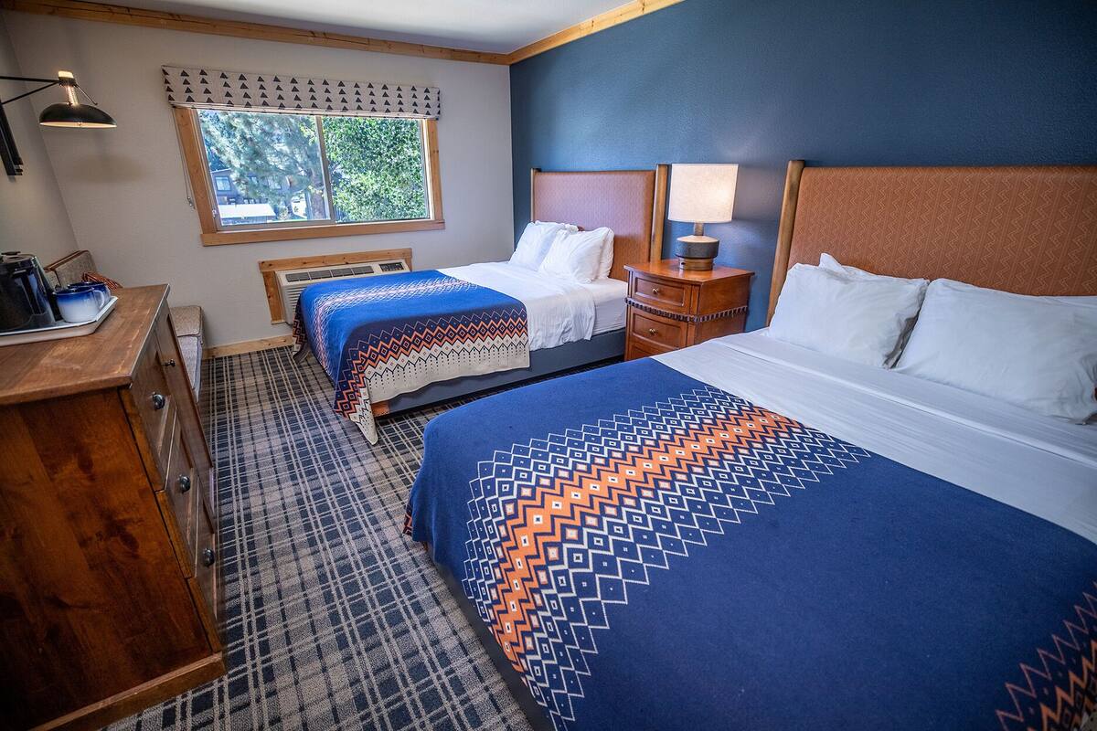 A hotel room with two beds, a wooden dresser, a desk with a lamp, and a window with a view of trees.