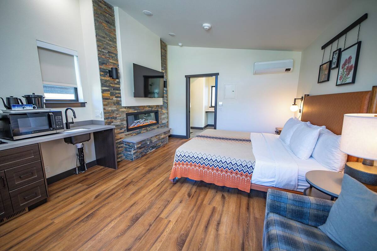 A hotel room with a kitchenette, a fireplace, a bed, a TV, and a bathroom.