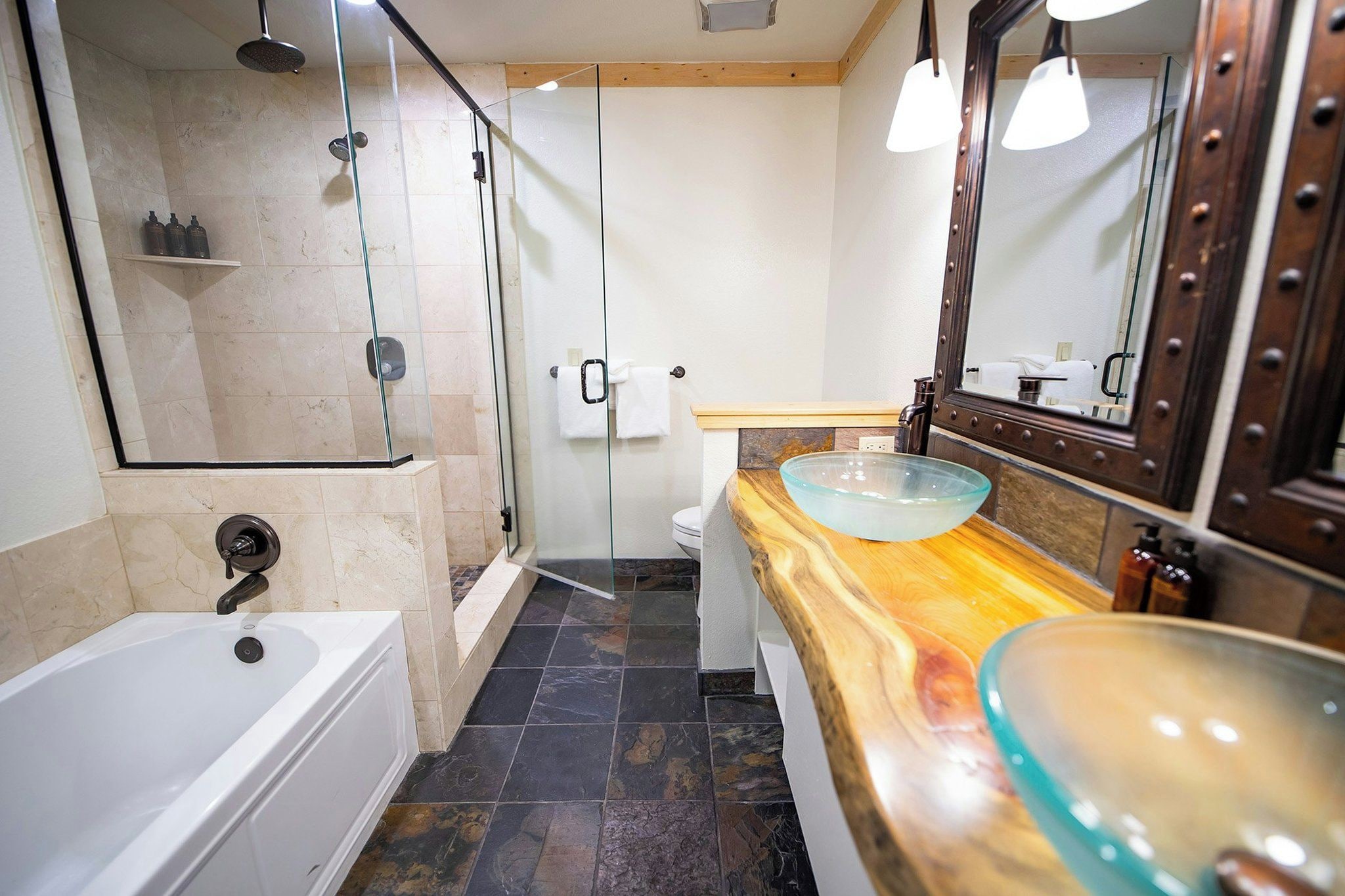 Un baño moderno con ducha envuelta en vidrio, una bañera blanca y un tocador de madera con lavabo de vidrio.