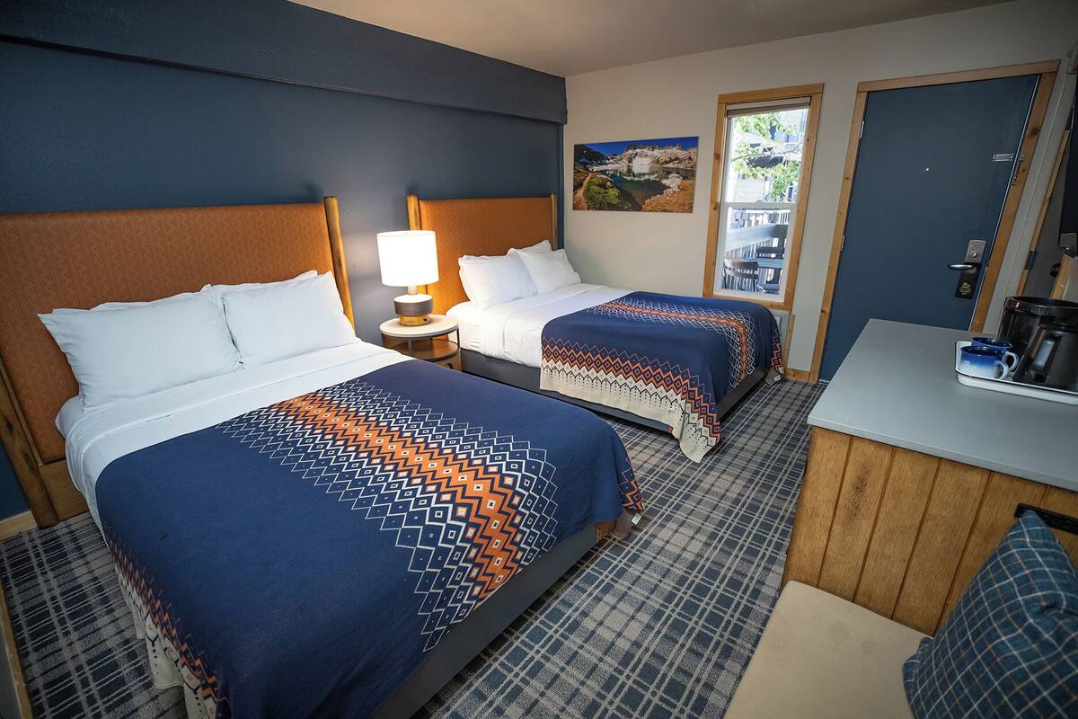 A hotel room with two beds, a small desk, a coffee maker, and a window with a view of a mountain and lake.