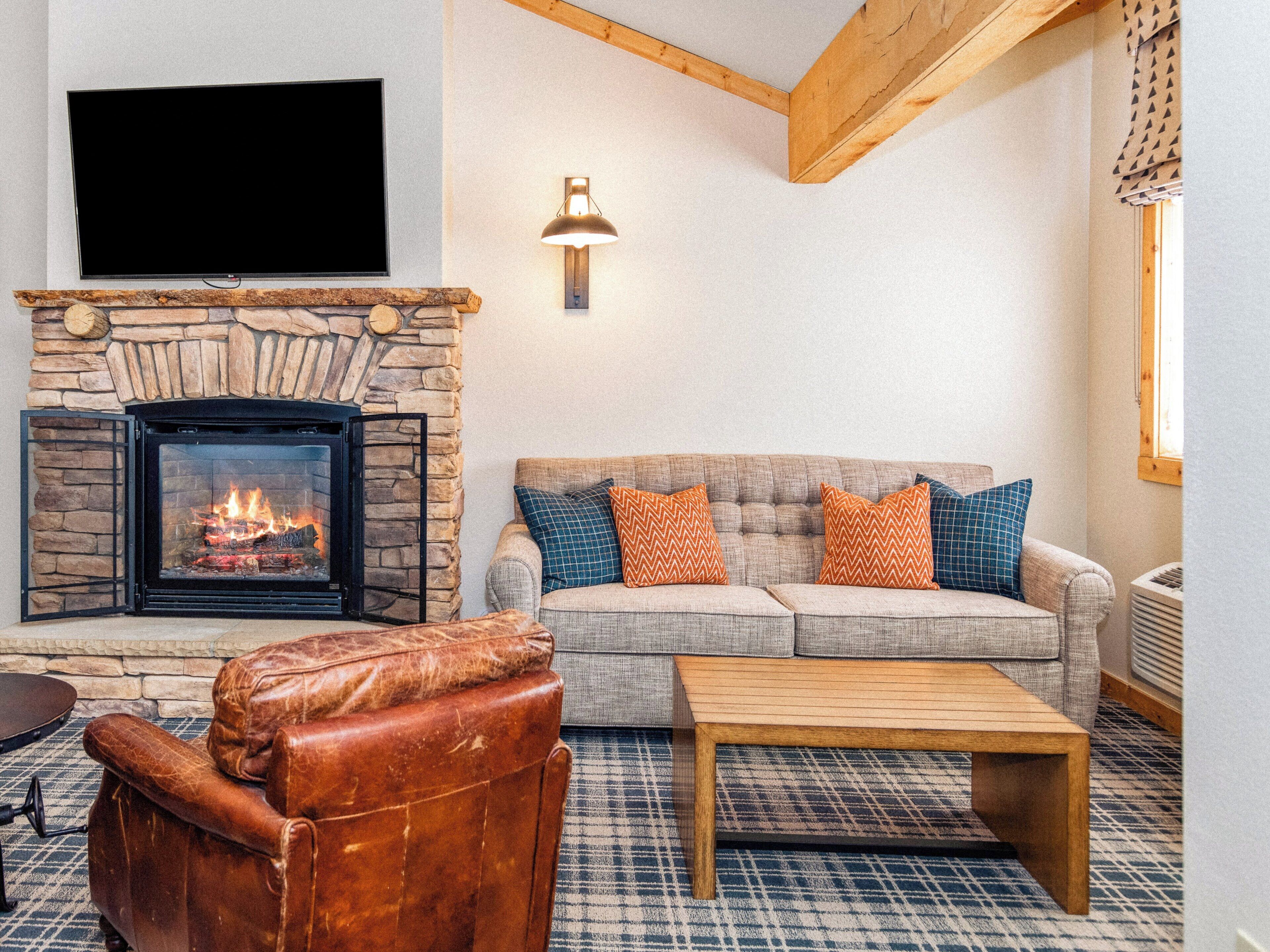 A living room with a stone fireplace, a television mounted on the wall, a sofa with patterned cushions, and a leather armchair.