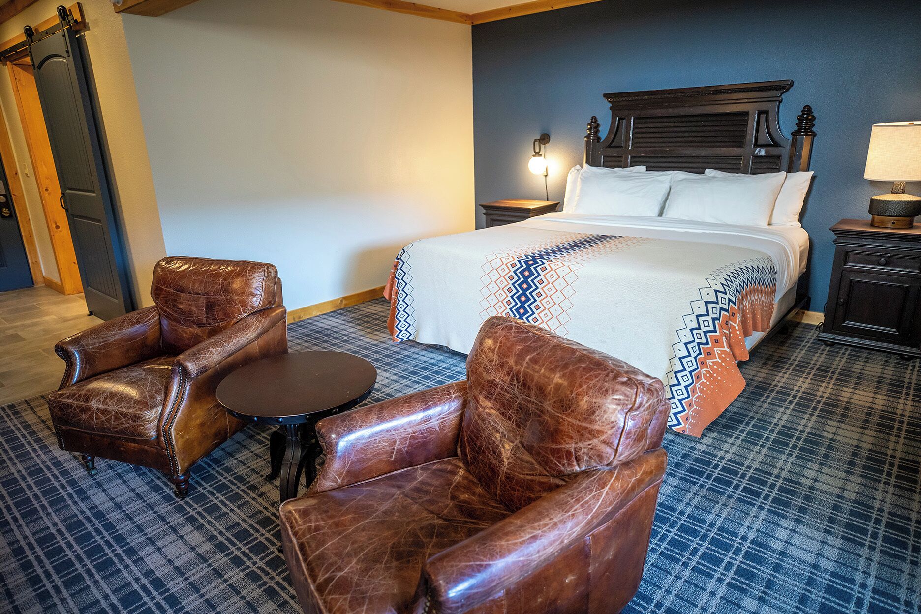 A hotel room with a large bed, two leather armchairs, a small round table, and a nightstand with a lamp.