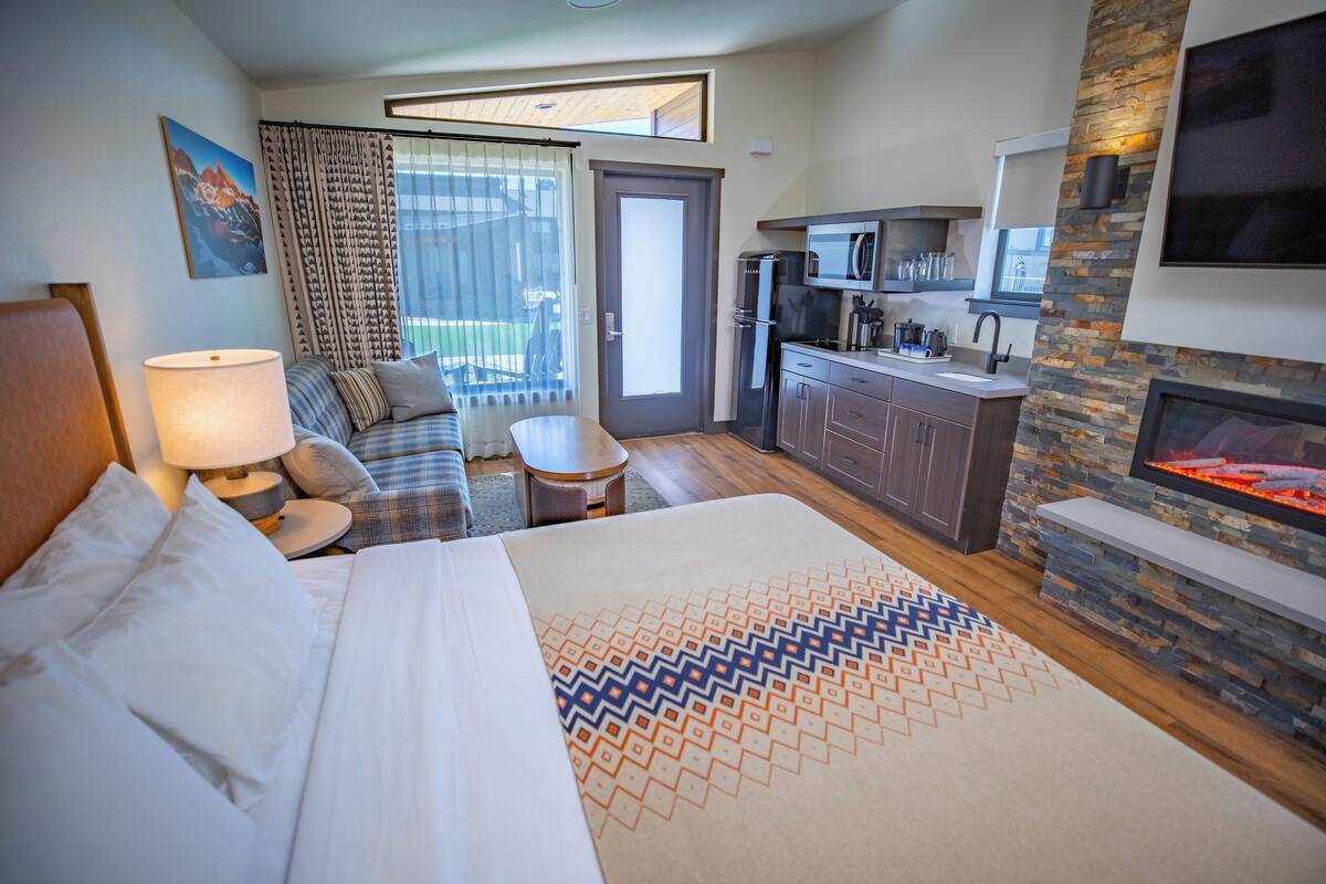 A hotel room with a bed, a sofa, a small table, a kitchen area, and a fireplace.