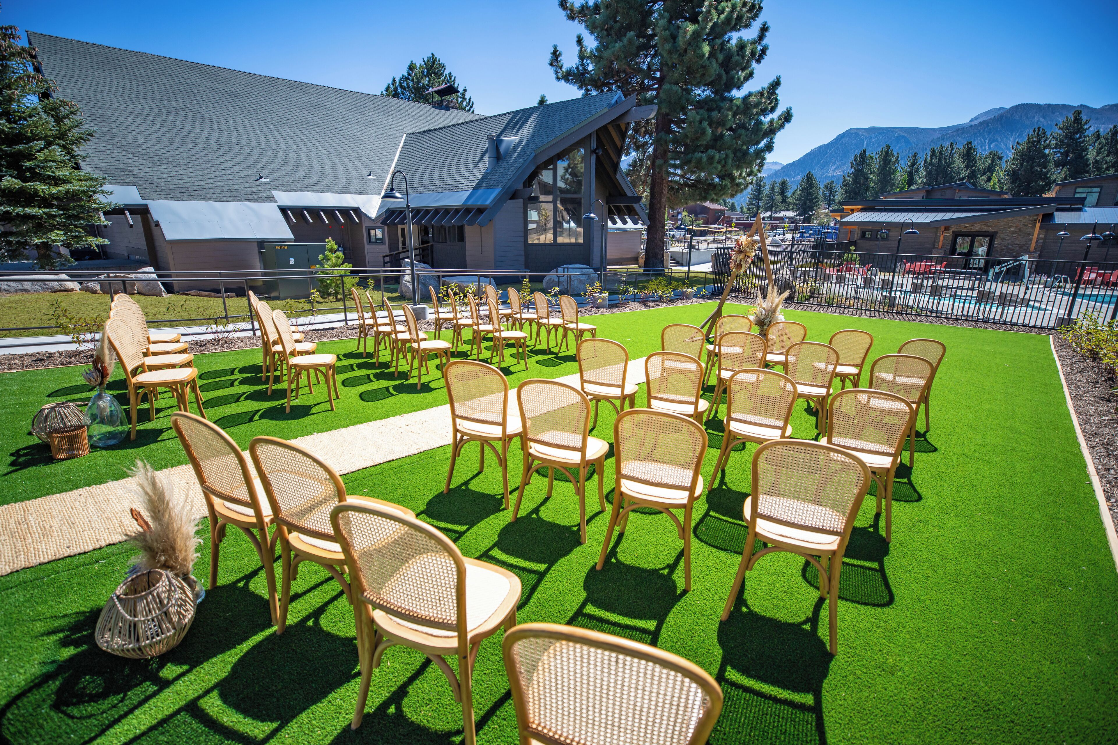 An outdoor seating area with wicker chairs arranged on artificial grass, a building with a sloped roof, and a mountain backdrop.