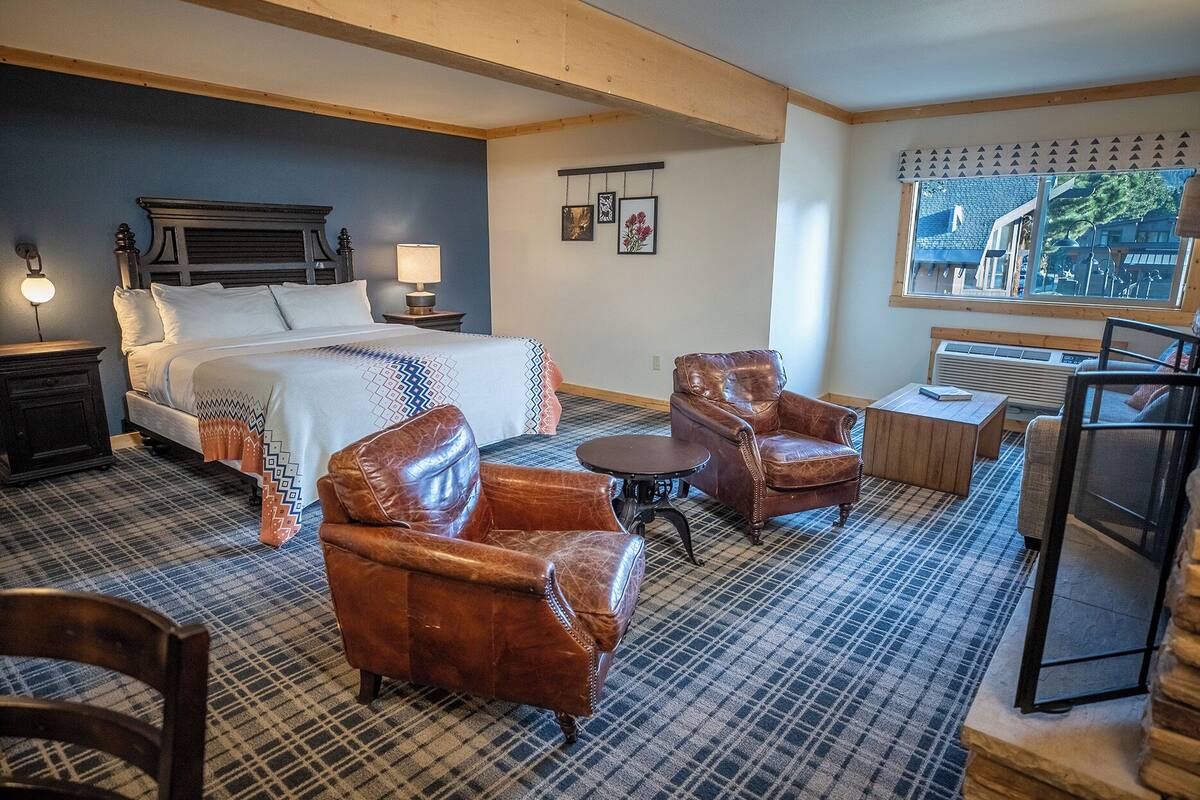 A hotel room with a bed, two leather armchairs, a small table, a nightstand, a lamp, a wall-mounted heater, and a window with a view of buildings.