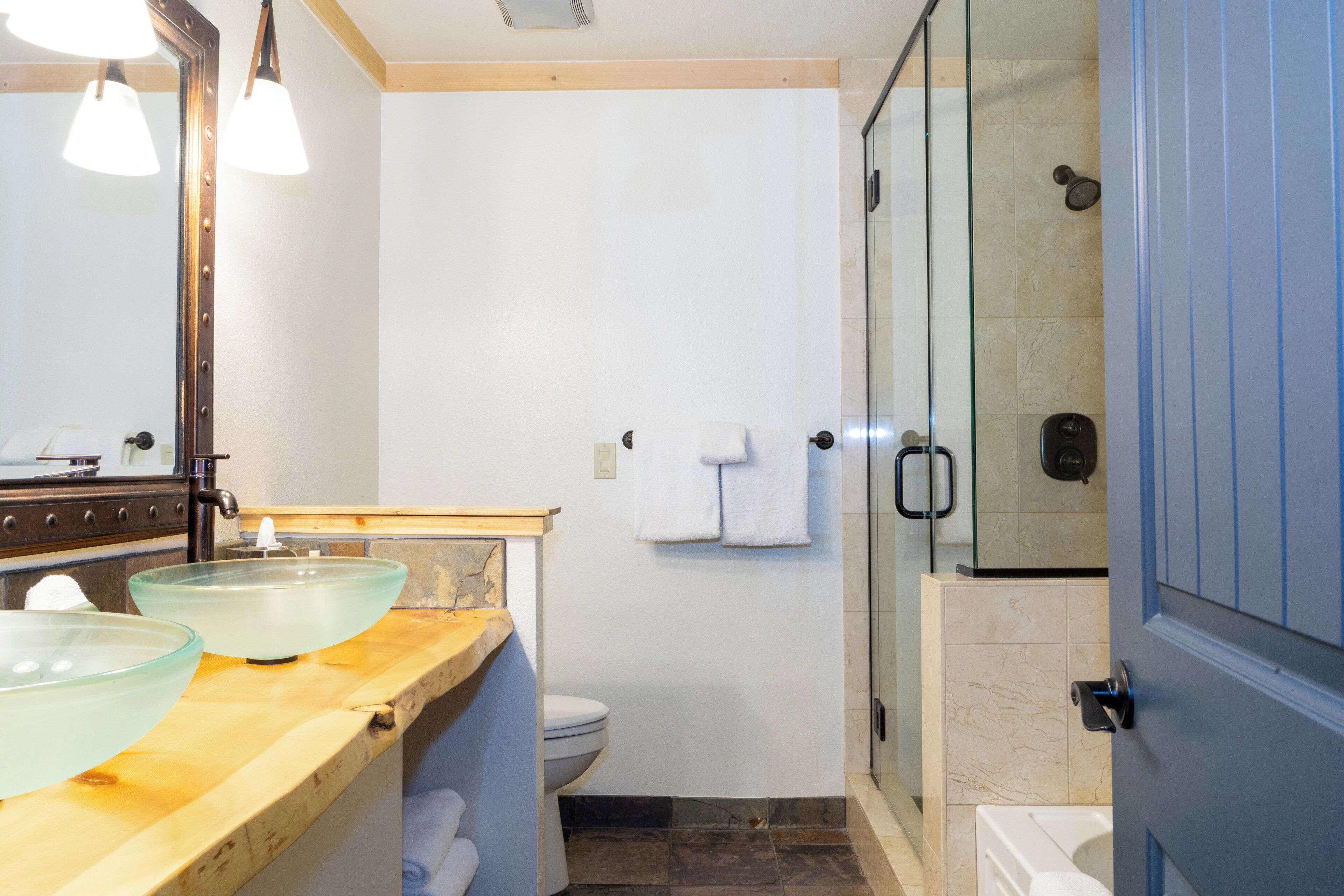 Une salle de bain avec un meuble-lavabo en bois, deux bols en verre, des toilettes, une cabine de douche en verre et une porte bleue.