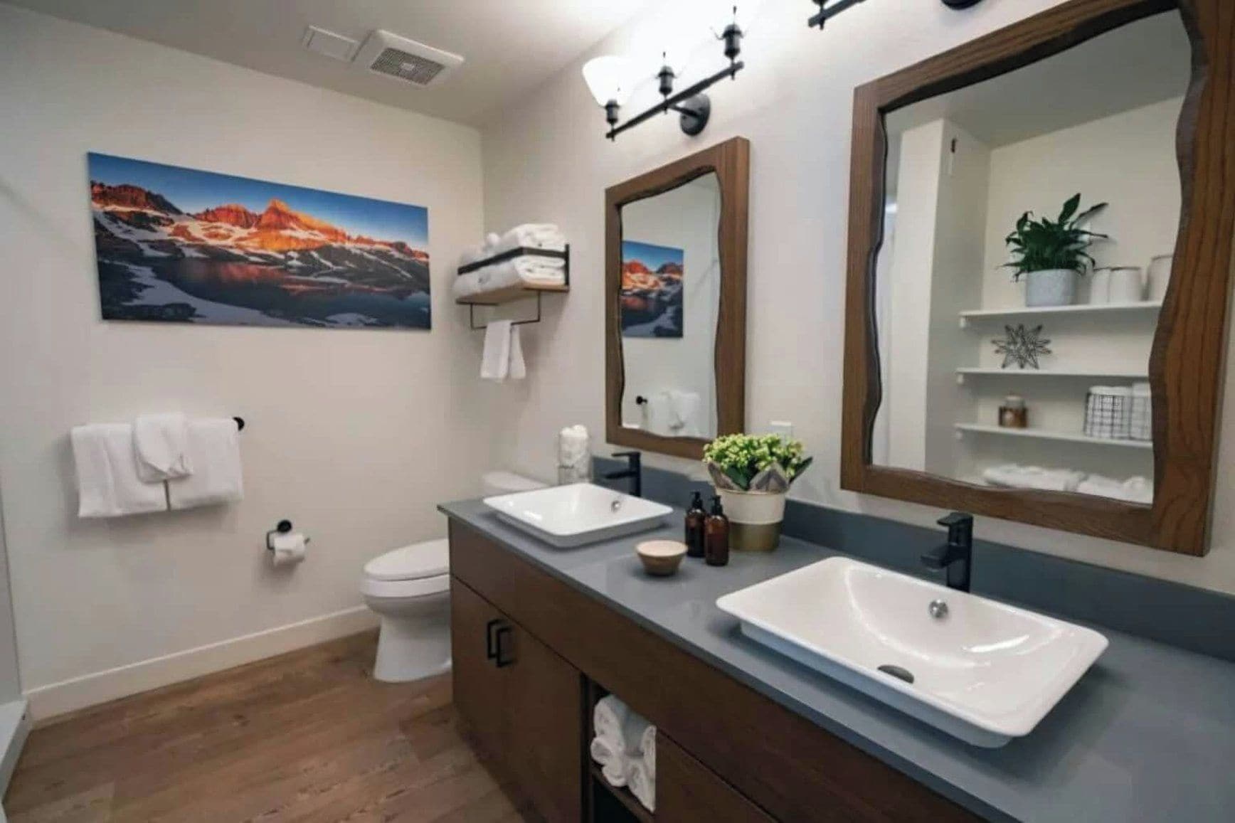 A modern bathroom with two sinks, a large mirror, and a painting of a mountain landscape.