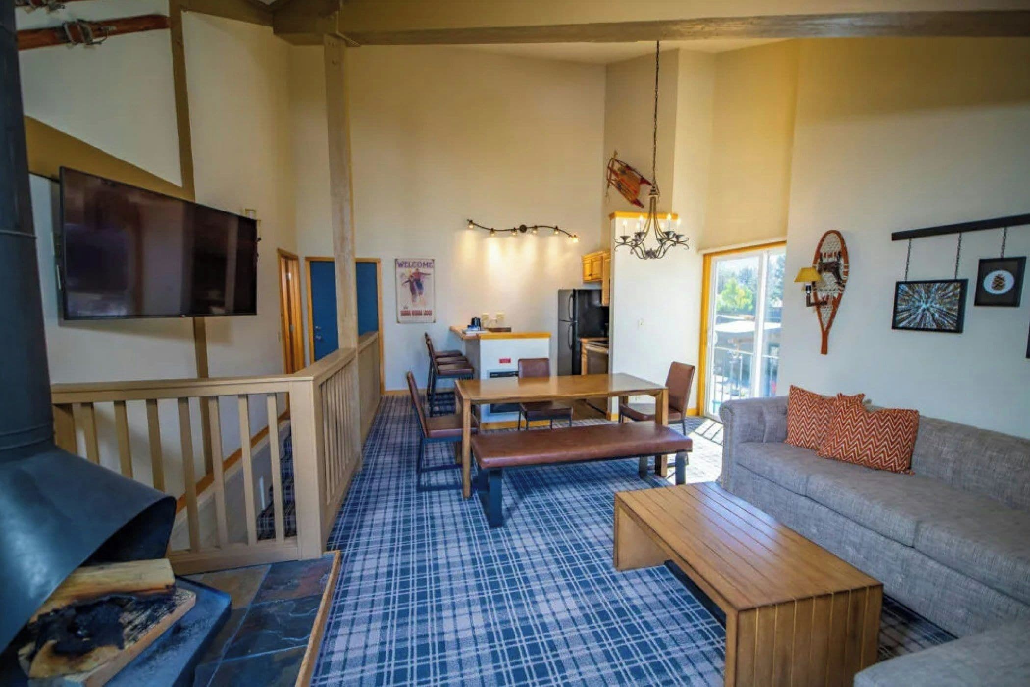 Un salon comprenant un canapé, un banc en bois, une table à manger avec des chaises, un téléviseur fixé au mur et un escalier.