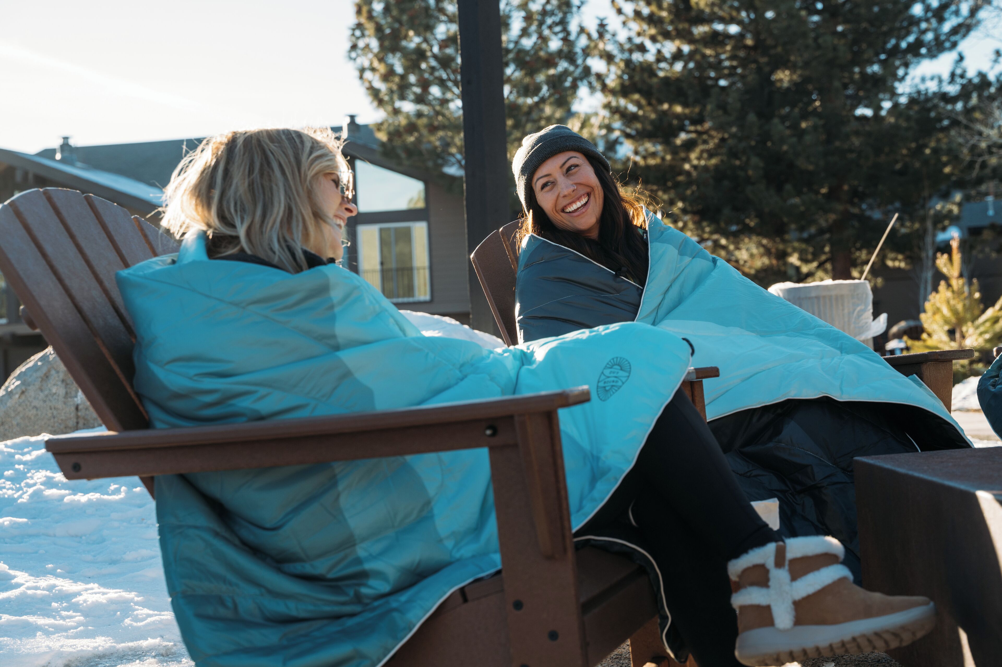 Two people sitting on a wooden deck chair outdoors with a house and trees in the background.