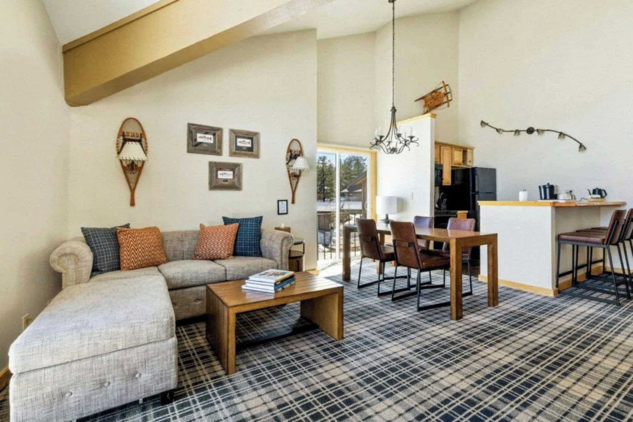 A living room with a sofa, coffee table, and dining area with chairs and a table. There is a kitchen visible in the background.