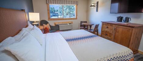 A hotel room with a bed, a desk, a chair, a TV, and a window with a view of trees.