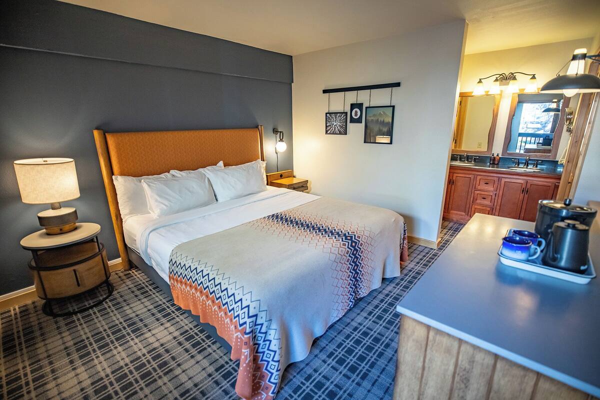 A hotel room with a bed, a nightstand, a lamp, a wall-mounted shelf with pictures, and a kitchenette area.