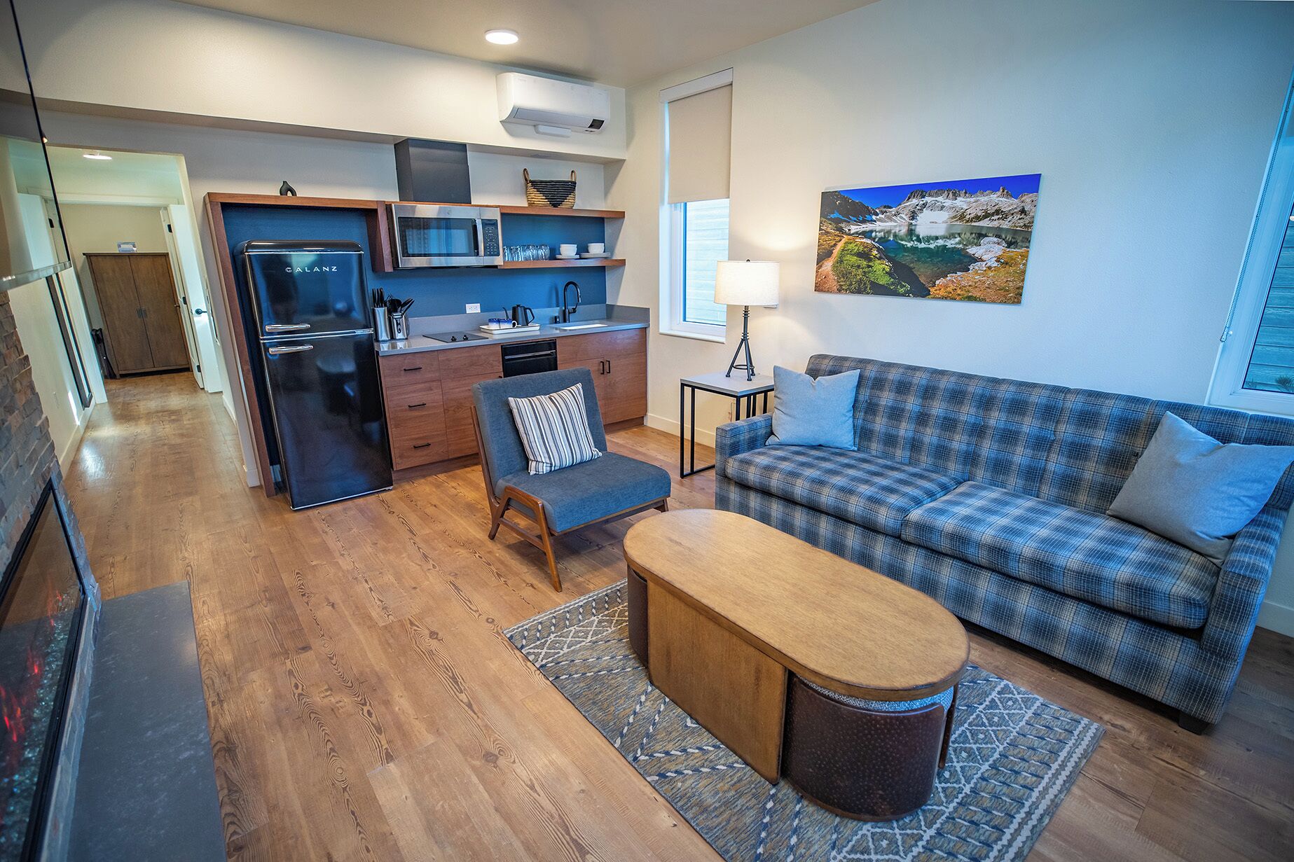 A modern living room with a sofa, a coffee table, and a kitchen area with a refrigerator, microwave, and sink.