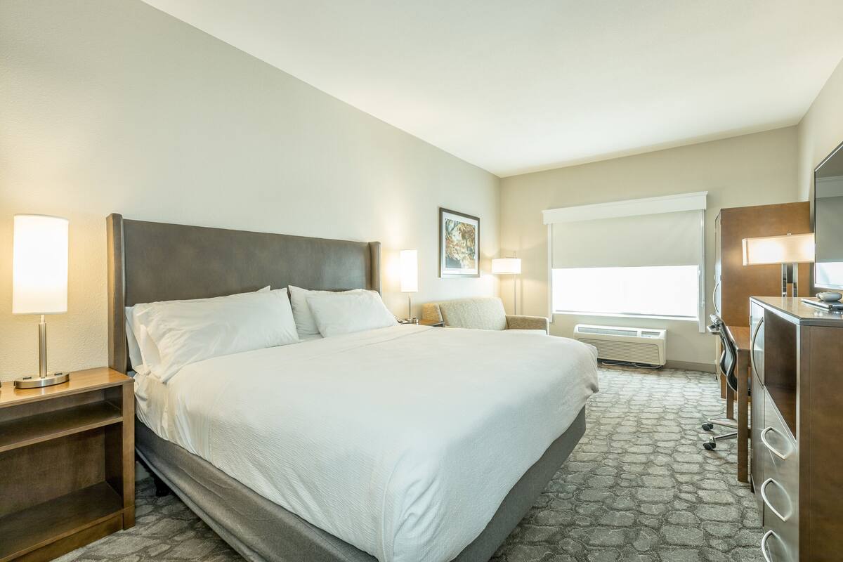 Une chambre d’hôtel avec un grand lit, des tables de chevet, un bureau avec une chaise, une fenêtre avec des stores et un téléviseur fixé au mur.