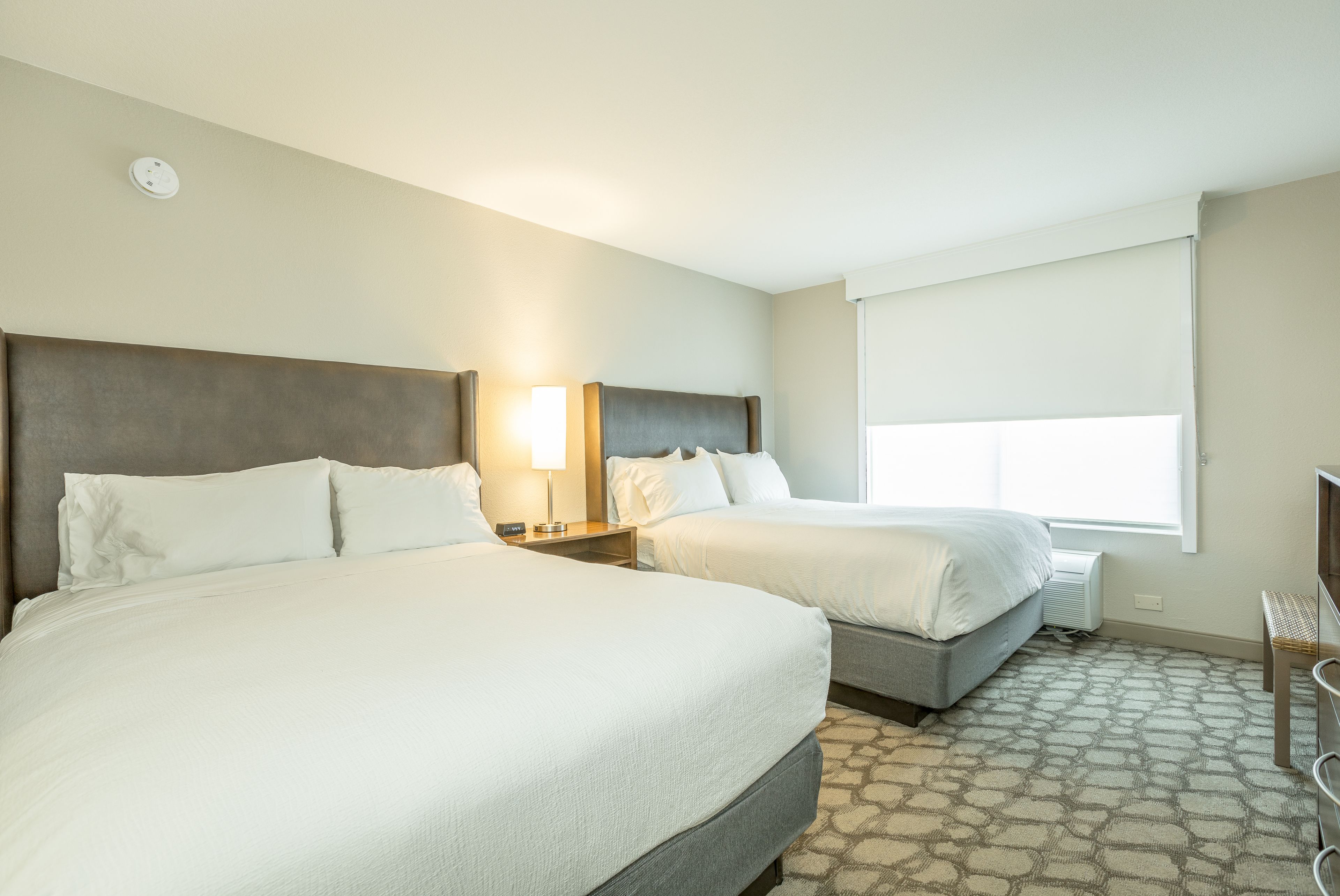 A hotel room with two beds, a nightstand with a lamp, a window with blinds, and a patterned carpet.