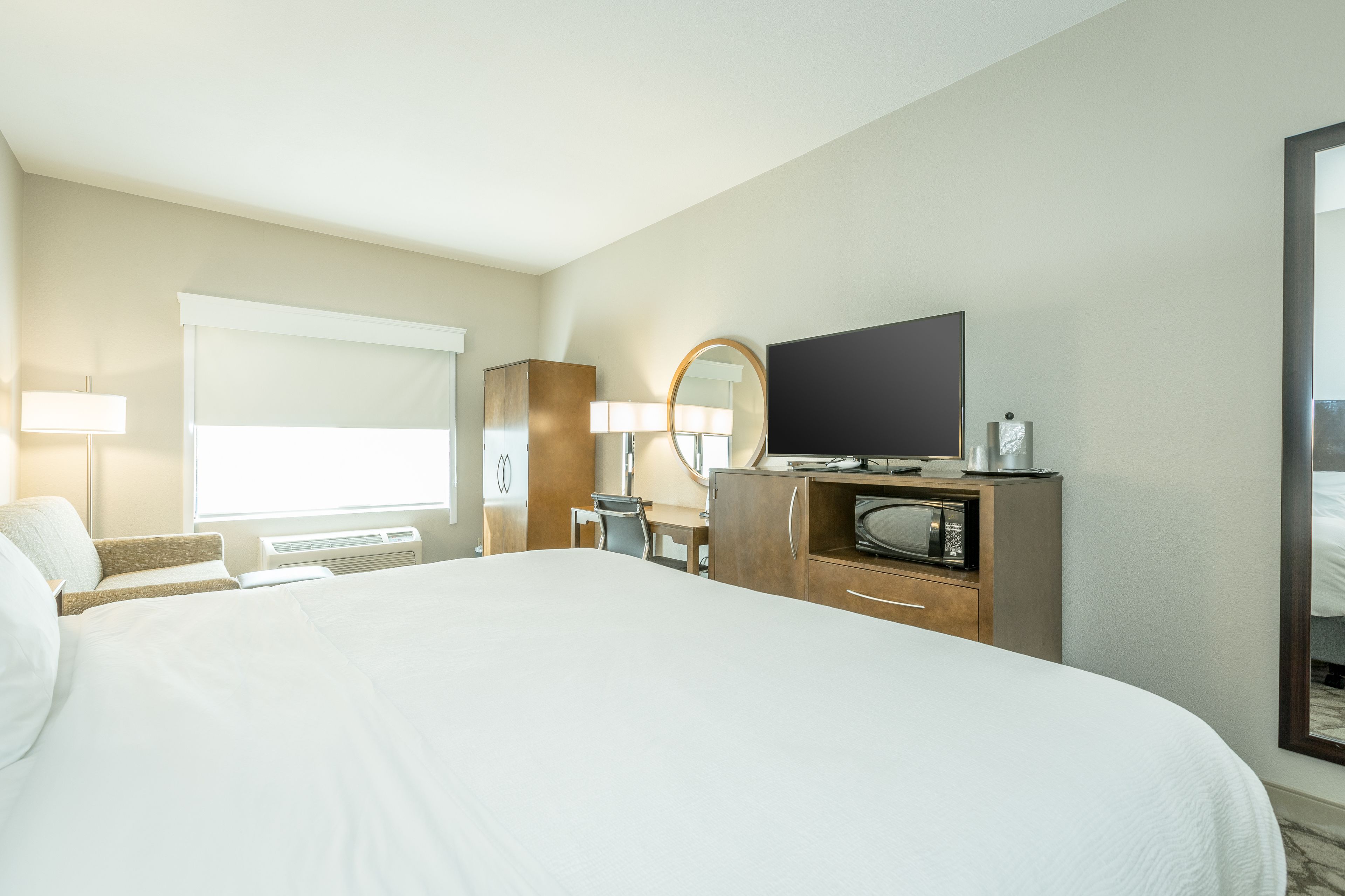 Habitación de hotel con una cama grande, un escritorio con silla, televisor, espejo y ventana con persianas.